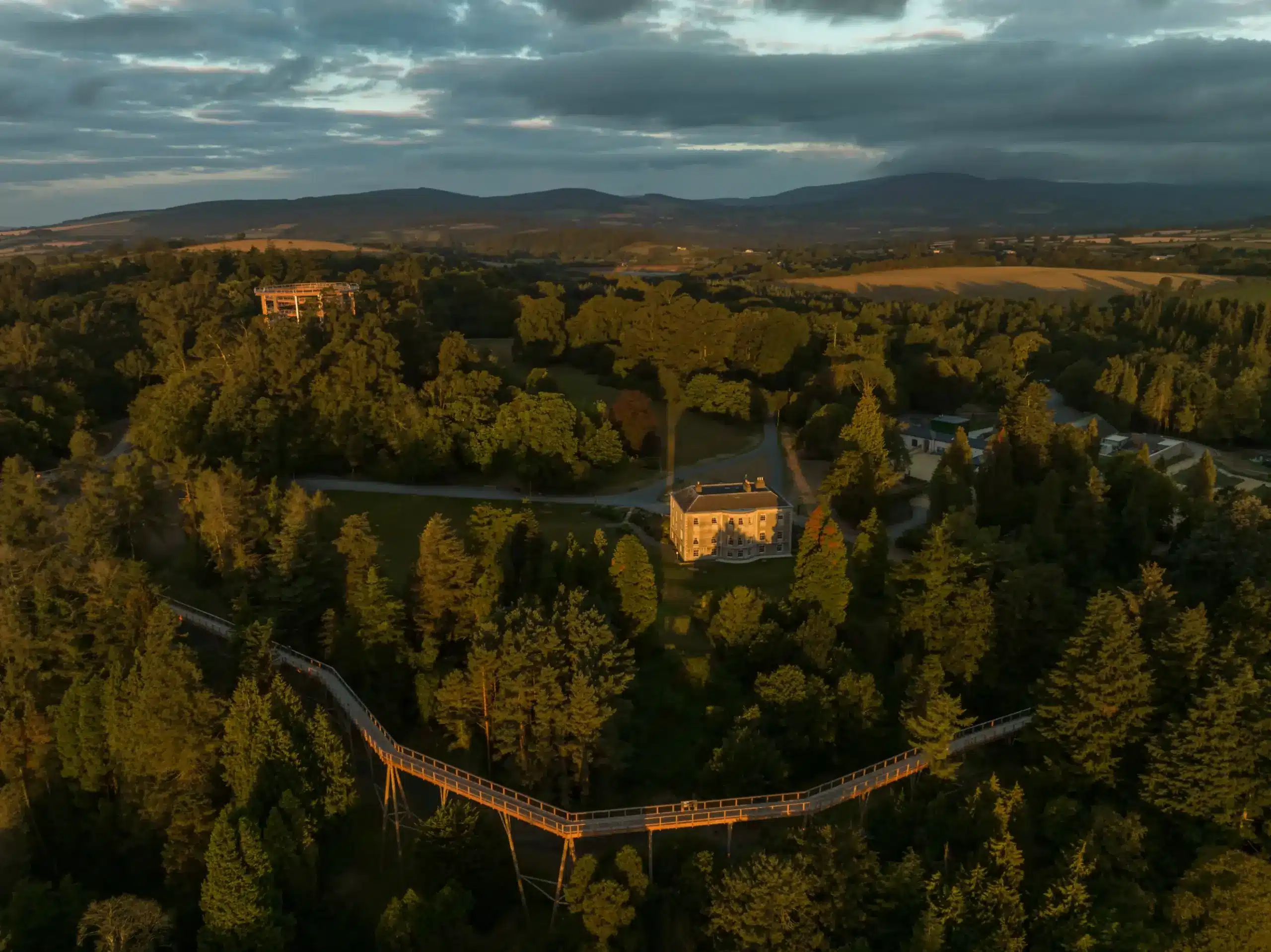 Aerial view of a treetop walkway winding through dense forest, leading to a large house. The scene is bathed in warm, golden sunlight with mountains and fields visible in the background.