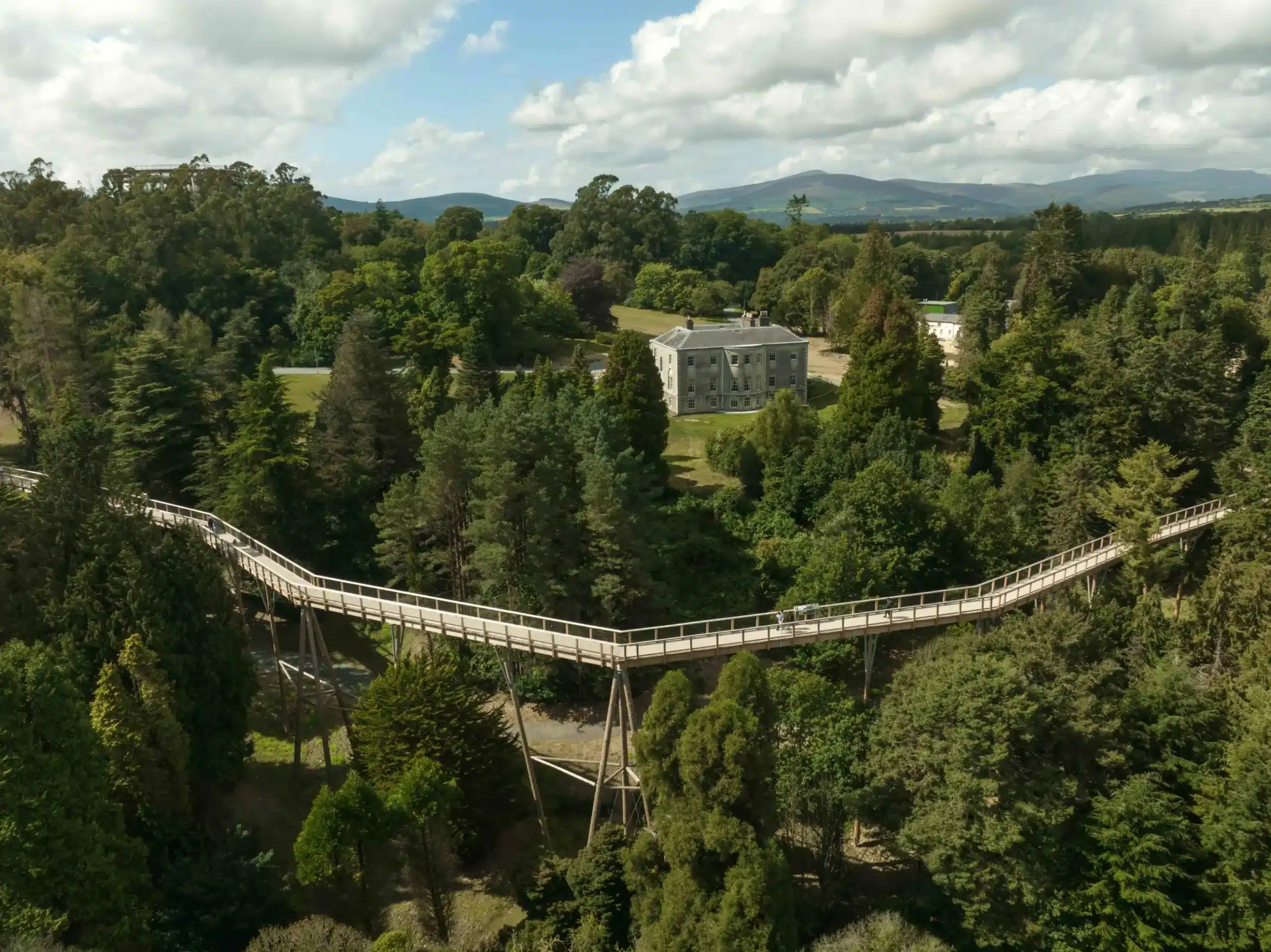 A long, elevated wooden walkway winds through dense green trees in a forest, with a large historic building in the background under a partly cloudy sky.