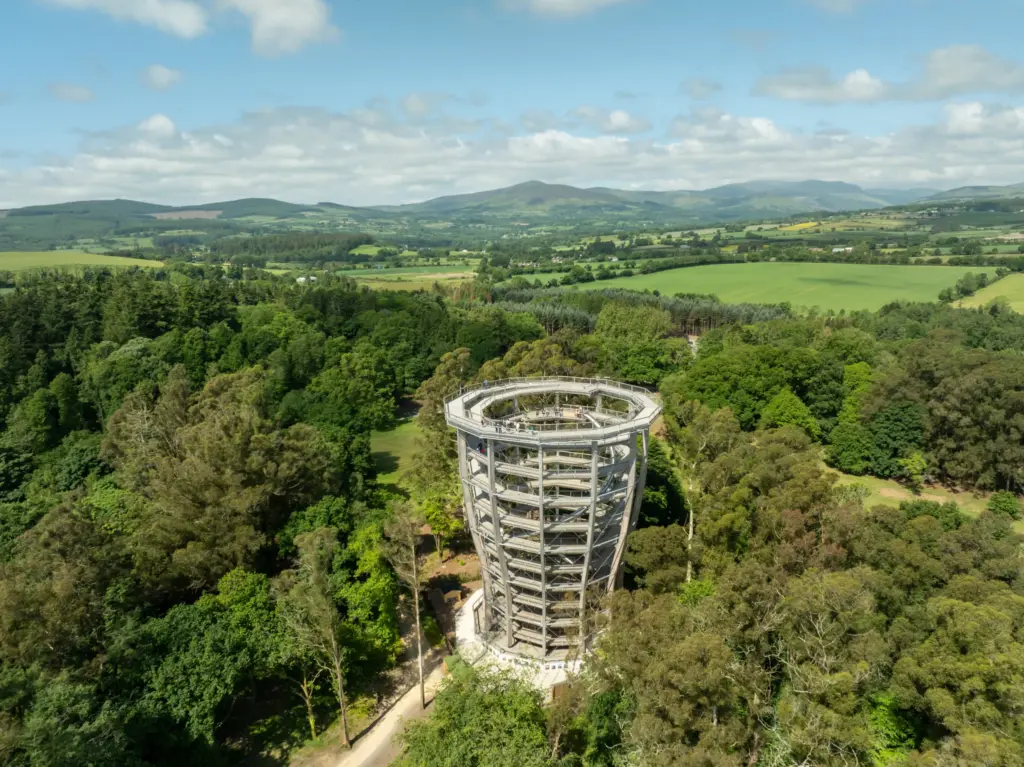 avondale - drone photos -34 1_2x A tall circular wooden observation tower rises above a lush green forest, surrounded by trees and fields, with rolling hills and a blue sky filled with clouds in the background.