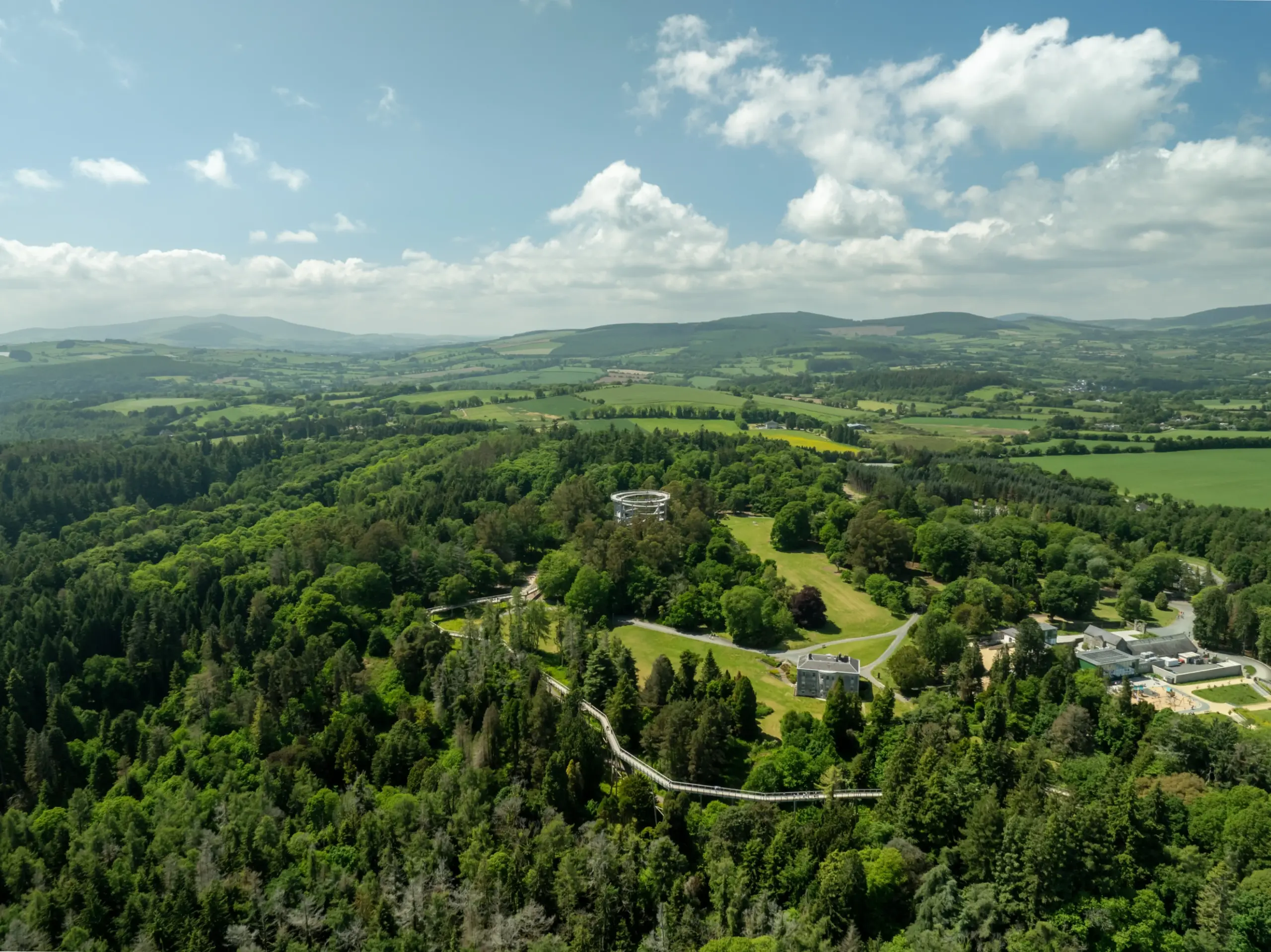 Aerial view of a lush green landscape with dense forests, open grassy areas, scattered buildings, and distant rolling hills under a blue sky with scattered clouds.