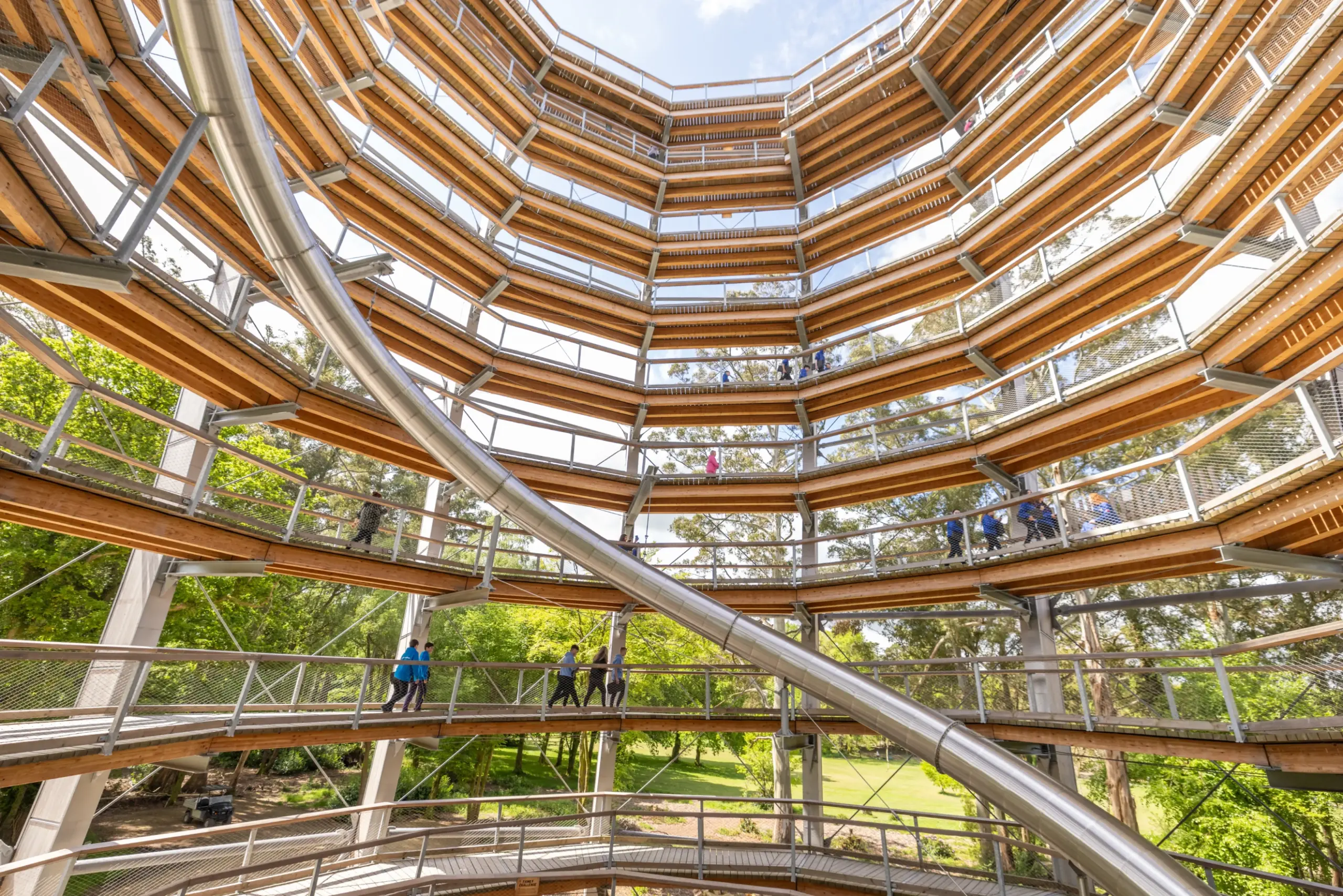 View from inside a tall, circular wooden observation tower with multiple levels, surrounded by trees, with people walking on spiral ramps and a large metal slide curving through the centre.