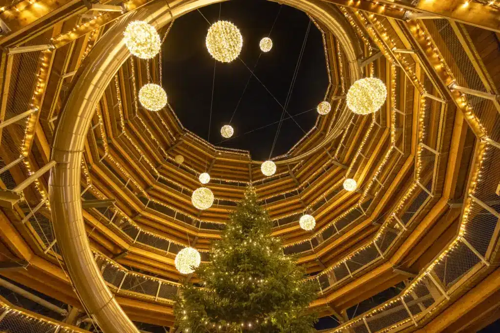 Avondale Evening Shoot - Camera -32 (1) Looking up from the base of a spiral wooden structure decorated with strings of warm yellow lights, glowing orbs, and a Christmas tree in the centre.