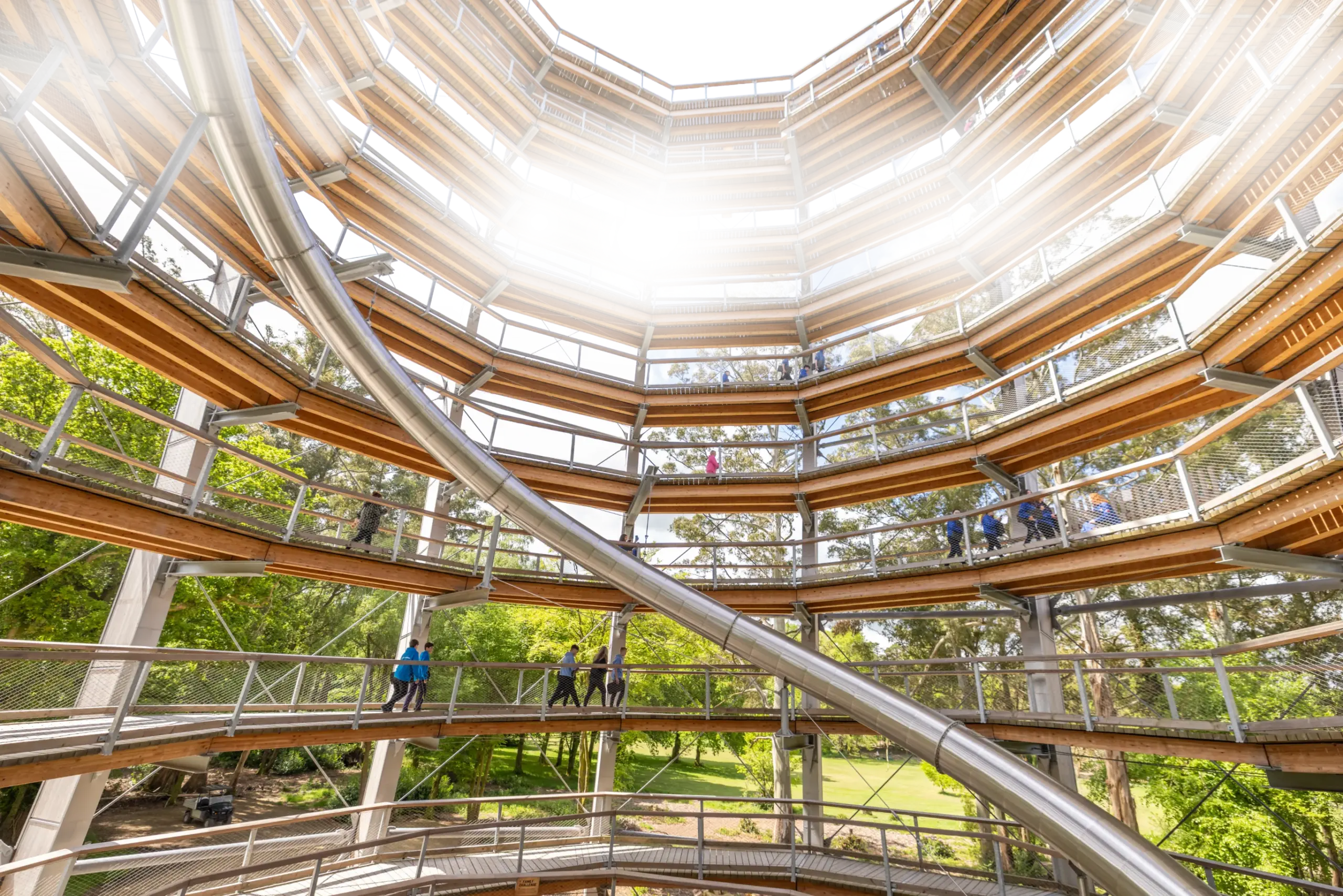 People walk inside a multi-storey, circular wooden structure with a spiral ramp and greenery visible outside. Sunlight streams in from the open top.