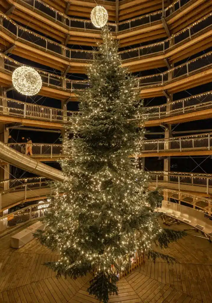 Christmas Lights - Camera-341 A large Christmas tree covered in warm white lights stands indoors under a high ceiling with balconies, decorated with more lights and glowing baubles.