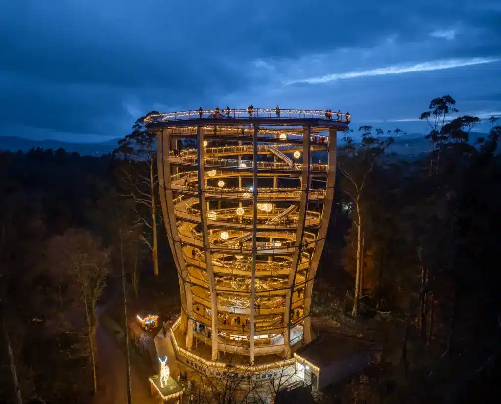 Christmas Lights - Drone28 A large, spiral walkway tower lit with warm lights stands among trees at dusk, with people visible on its ramps and top platform. The evening sky is deep blue and mountains are in the distance.