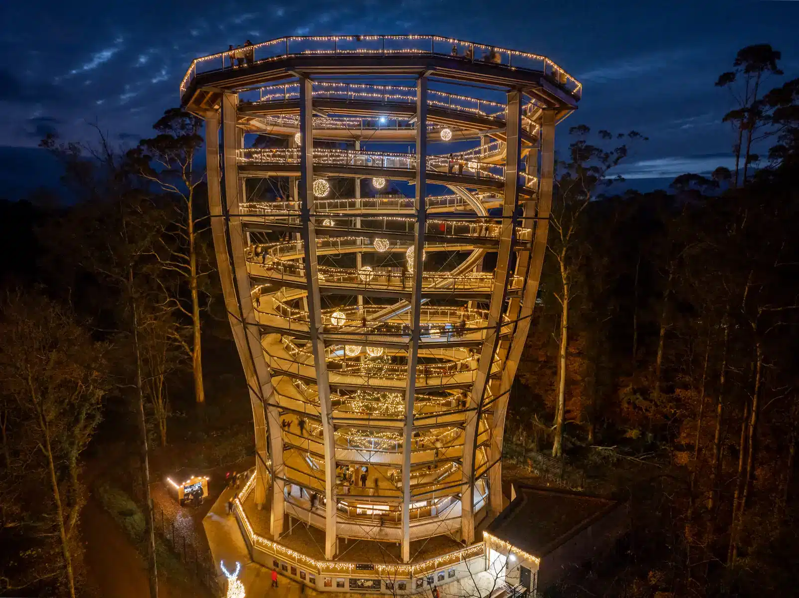 A large, illuminated wooden observation tower spirals upward through a forest at dusk, with people walking along its paths and festive lights decorating the structure.