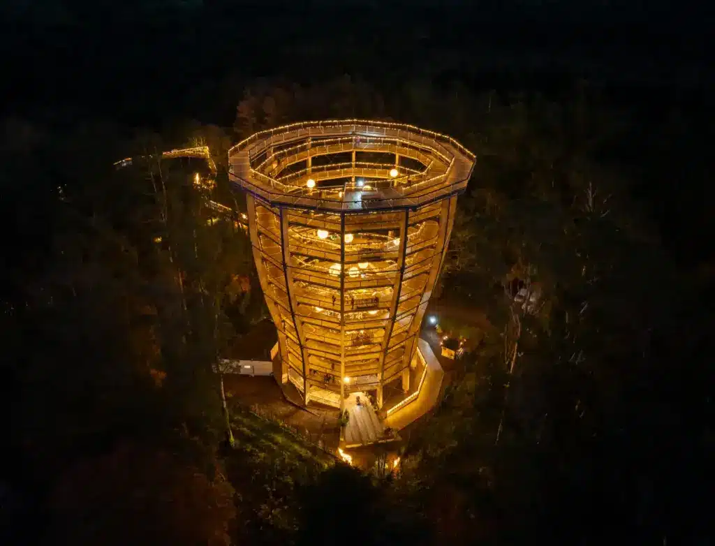 Christmas Lights - Drone49 A tall, spiral wooden tower illuminated by warm lights at night, surrounded by trees in a forested area.