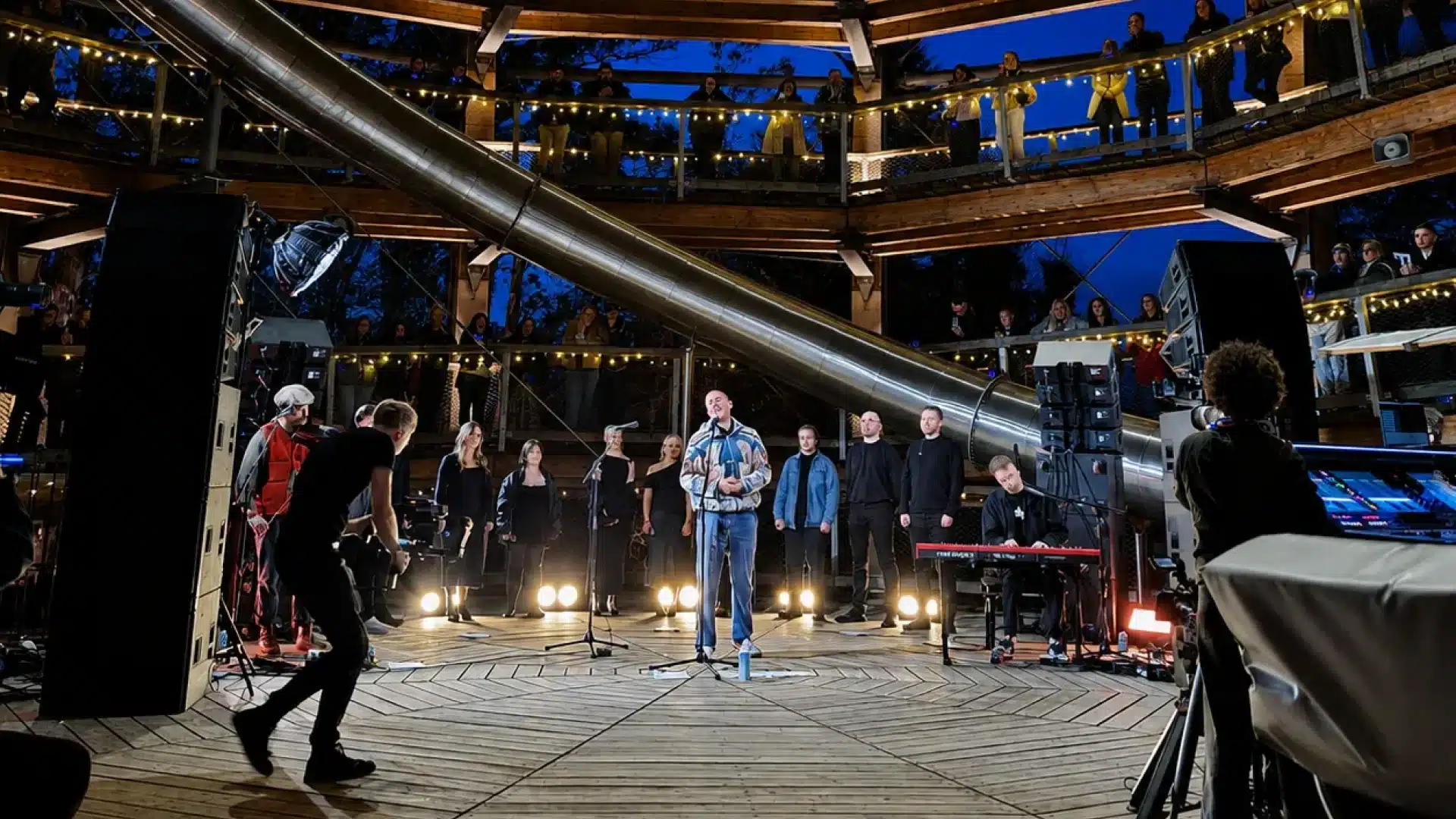 A singer performs on a circular wooden stage at night, surrounded by musicians and vocalists. People watch from multiple levels of a spiral wooden structure with lights. A photographer captures the scene.
