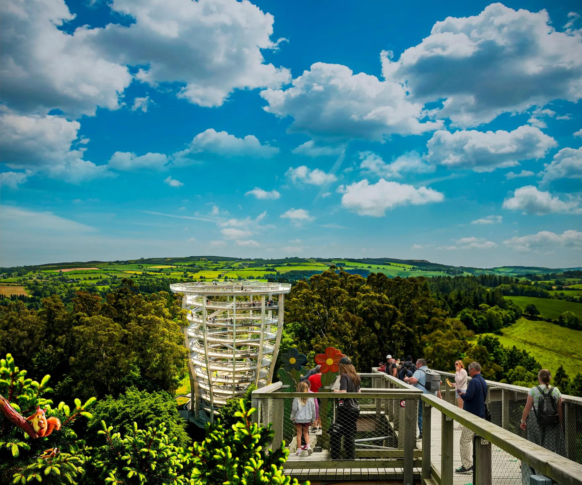 People stand on a wooden viewing platform overlooking lush green hills and forests under a bright blue sky with scattered clouds. A spiral-shaped observation structure rises from the trees.