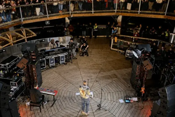 dermot kennedy2990 1_1x A performer stands at the centre of a round wooden stage, surrounded by speakers, instruments, and an audience watching from multiple levels in a circular indoor venue.
