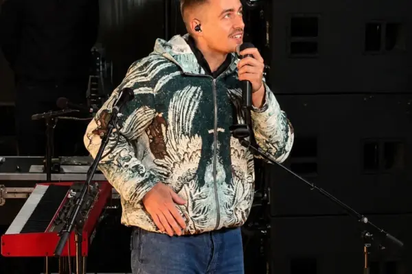 dermot kennedy73848 1_1x A man in a patterned hoodie and jeans holds a microphone onstage, standing near a keyboard. Another man in black stands behind him. Large speakers and lighting equipment are visible in the background.