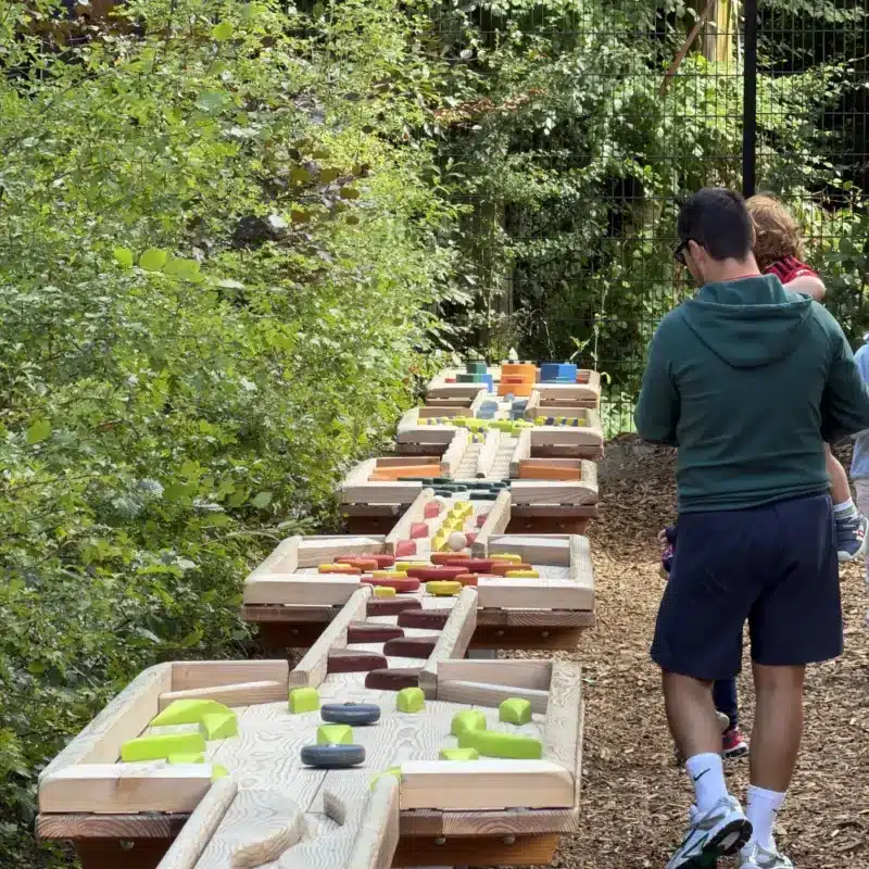 People walk along a path beside colourful interactive wooden structures designed for play, surrounded by greenery and trees in an outdoor setting.