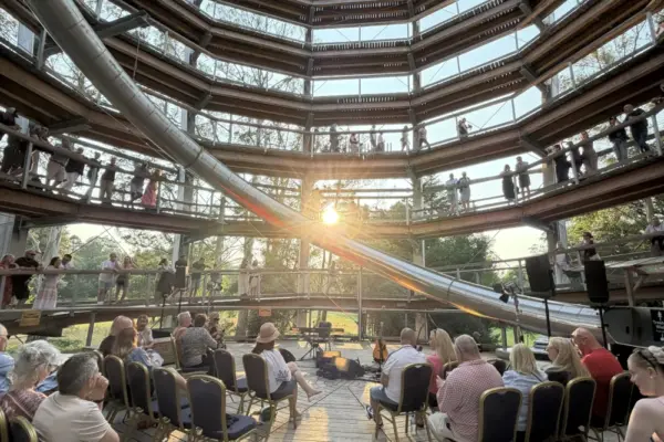 paddy casey_1675 1_1x People sit on chairs in a circular wooden structure with several levels. Sunlight streams through the centre as others stand or walk along railings above, and a long slide winds down from the upper floors.