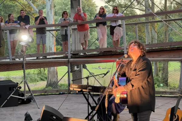 paddy casey_1705 1_1x A musician sings and plays guitar on an outdoor stage at sunset, with people watching from two levels of a wooden, circular structure surrounded by trees.