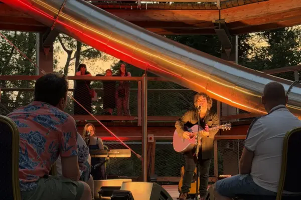 paddy casey_1732 1_1x A musician plays guitar and sings on an outdoor stage while people watch from chairs and upper floors. A large metal slide and wooden structure are visible, with warm evening light illuminating the scene.