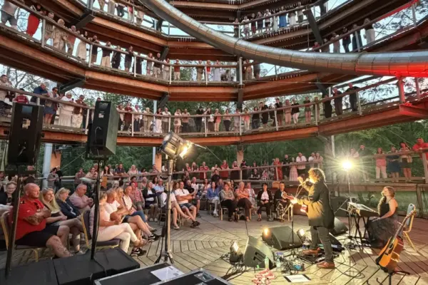 paddy casey_1761 1_1x A musical performance takes place at the base of a multi-level wooden structure, with seated and standing audience members watching from various elevated walkways. Instruments, lights, and sound equipment are visible on the stage.