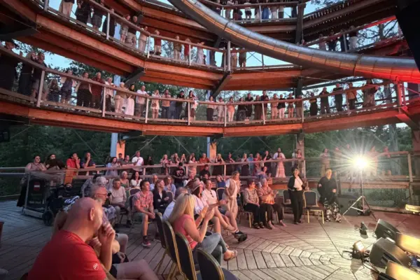 paddy casey_1788 1_1x A large audience sits and stands on multiple levels of a circular wooden structure, watching a performance on a stage with bright lights. Some people are seated on chairs, whilst others lean on railings above.