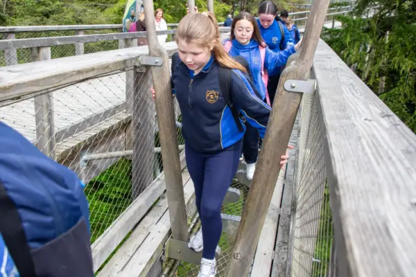 School-Priamry-013 1_2x A group of children in school uniforms walk carefully across a wooden and metal bridge outdoors, surrounded by trees, during what appears to be a school trip or outdoor activity.