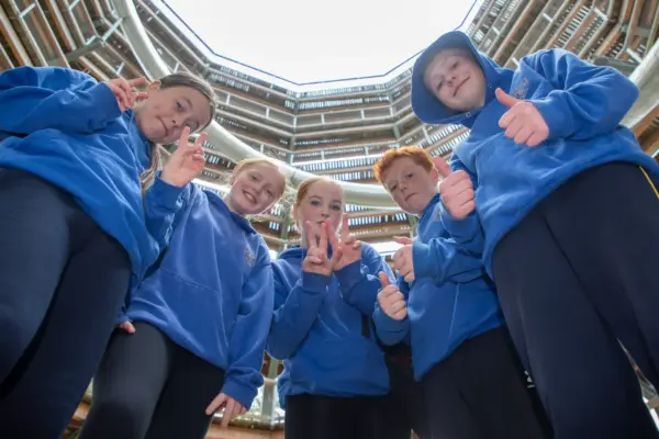 School-Primary-011 1_2x Five kids in blue hoodies stand in a circle, looking down at the camera and giving thumbs up, with a tall, circular building structure seen above them.
