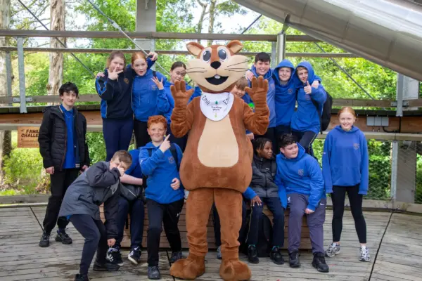 School-Primary-012 1_2x A group of kids in blue uniforms pose and smile around a person in a brown otter mascot costume on a wooden platform outdoors with trees in the background.