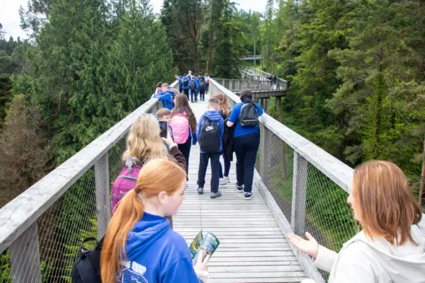 School-Primary-014 1_2x A group of people, mostly students with rucksacks, walk along a wooden walkway suspended above a forest, surrounded by tall green trees under a cloudy sky.