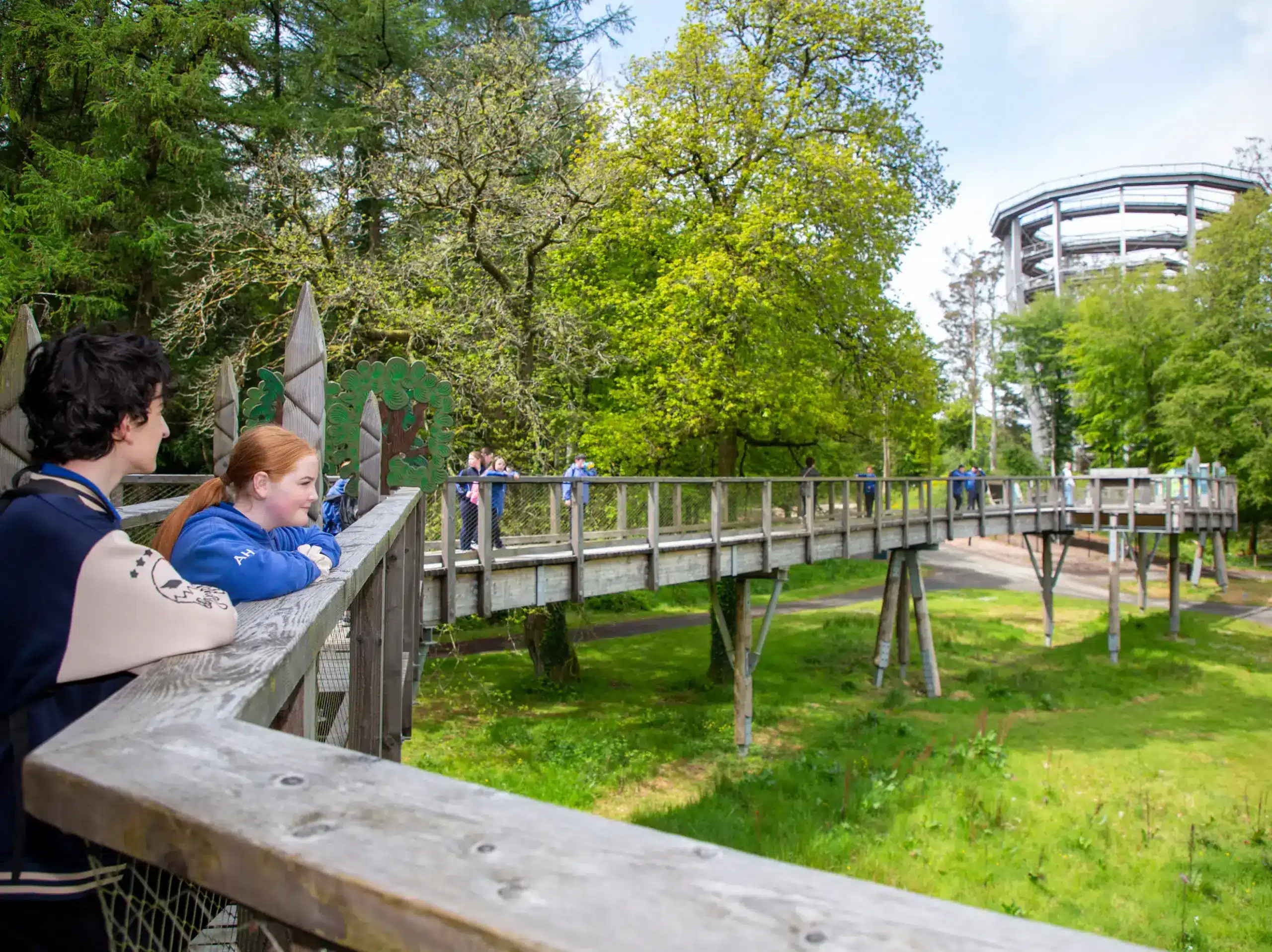 Two people stand on a wooden walkway overlooking a green forest area with more people walking in the distance. A tall circular structure is visible among the trees.