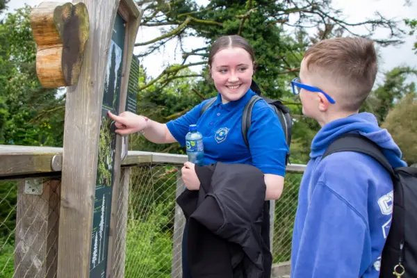 UL3A8845 1_2x A girl in a blue shirt points at an outdoor information board whilst smiling at a boy in glasses. They are standing on a wooden walkway surrounded by trees.