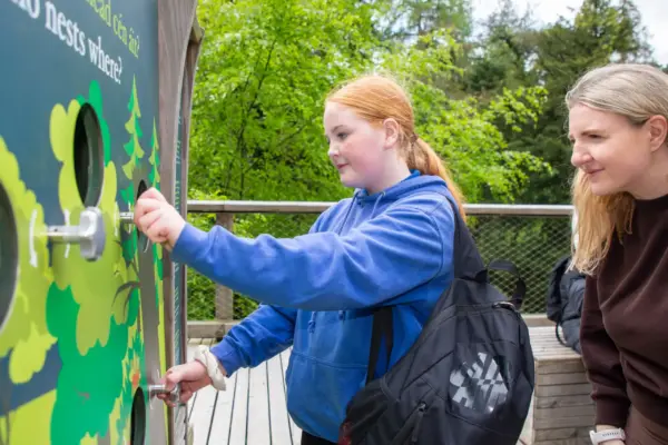UL3A8849 1_2x A girl in a blue hoodie interacts with an outdoor educational display as a woman watches. Trees and greenery are visible in the background.
