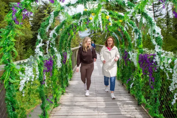 UL3A8873 1_2x Two women walk on a wooden bridge decorated with green leaves, white, and purple flowers, surrounded by trees. Both are smiling and enjoying the outdoor setting.