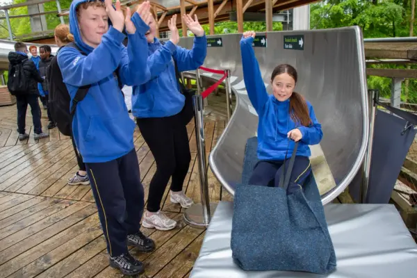 UL3A8916 1_2x Three children in blue hoodies, two clapping and cheering, watch as a girl exits a metal slide onto a padded mat, holding a large fabric sack, on a wooden decking outdoors.