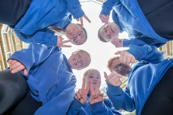 UL3A8919 1_2x Five people in blue hoodies stand in a circle, looking down at the camera from below, smiling and making peace signs and thumbs-up gestures. The bright sky and parts of a building are visible above them.