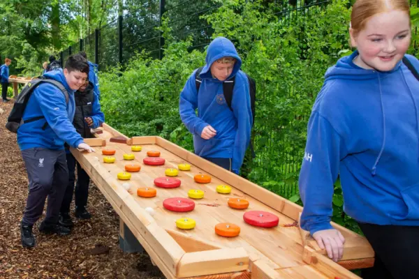 UL3A8974 1_2x Four children in blue hoodies play a large outdoor wooden board game with red and yellow discs in a wooded area, smiling and enjoying themselves.